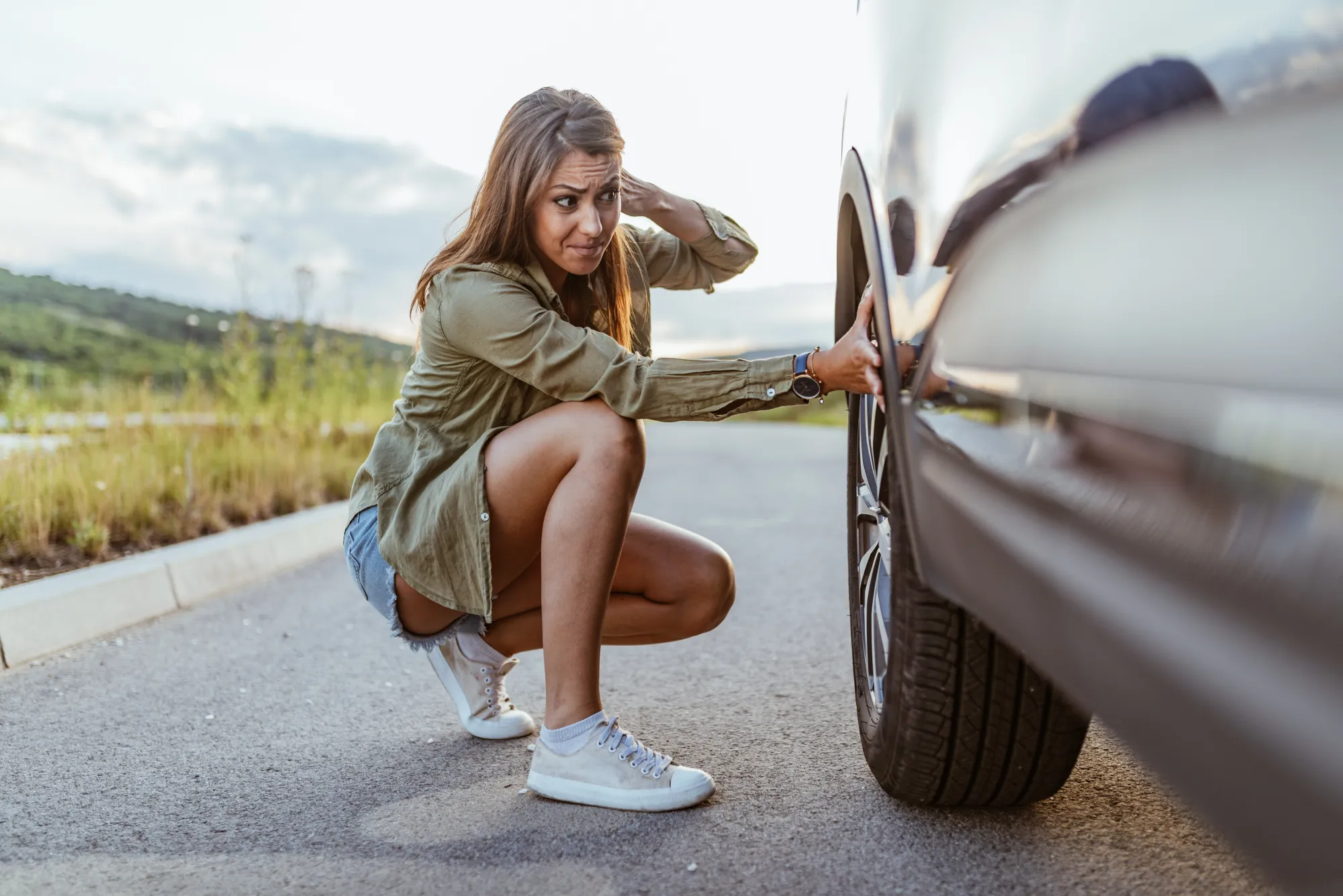 A worried woman crouches next to the front tire of her car on the side of a road.